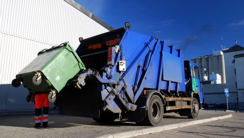 A large blue and black rubbish collection truck is positioned on a paved area in an urban setting, with its rear hopper open, allowing the loading of waste materials. The truck's rear compactor mechanism is visible, with metal components and hydraulic arms engaged in lifting and compressing waste. A green wheeled waste bin is being emptied into the truck by a worker wearing high-visibility orange trousers, though only the lower half of the worker is visible. The background shows a modern white industrial building with metallic siding and a clear blue sky above. The scene is well-lit, indicating daylight, and the environment appears clean and organized, reflecting a professional rubbish removal service in the Richmond area.
