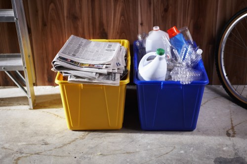 Two large plastic recycling bins positioned on a concrete floor in a garage or storage area, with a wooden wall in the background. The bin on the left is yellow and contains a stack of newspapers and assorted paper waste, while the bin on the right is blue and filled with various plastic bottles, some with caps, and other plastic packaging materials. Part of a bicycle wheel is visible on the right side of the image. The scene suggests a household or small-scale rubbish collection setup, typical of home or office clearance services in Richmond, highlighting the segregation of paper and plastics for recycling.