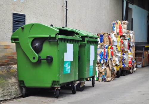 A large, green wheeled rubbish bin made of plastic with a rounded lid and a black wheel on the side, positioned on a grey paved surface next to a concrete and brick wall; to the right, there are multiple tightly packed bundles of flattened cardboard boxes with various colours and printed markings, stacked against a building with a glass window and a metal structure, suggesting an outdoor storage area in an urban or commercial environment, typical of a rubbish removal service in Richmond targeting household or office waste. The scene is illuminated by natural light, with clear details of the bin's textured surface and the corrugated cardboard's folds and branding visible. This setting reflects the typical waste collection and clearance situation handled by Office Clearance Richmond in the local postcode area.