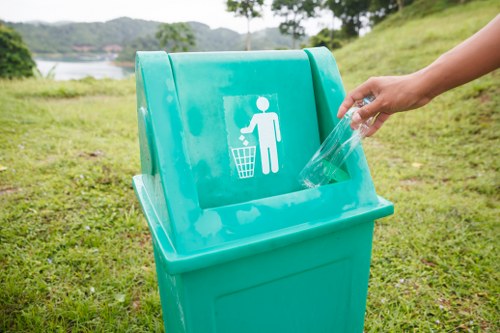 A person is discarding a clear plastic bottle into a bright turquoise outdoor trash bin with a white icon of a figure throwing waste into a bin, situated on a grassy area near a body of water and green hills. The environment appears clean and natural, with the bin positioned on a well-maintained lawn and the scene suggesting an emphasis on waste disposal and recycling practices. The image highlights proper rubbish management in a scenic outdoor setting, aligning with services offered by Office Clearance Richmond in Richmond, London.