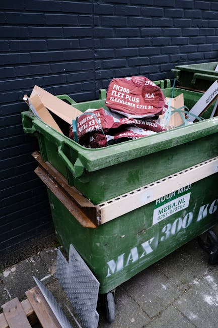 A green plastic waste bin positioned on a paved outdoor area adjacent to a dark brick wall. The bin is filled with red bags of garden or landscaping materials, including bags labeled 'FK200 Flexy Plus