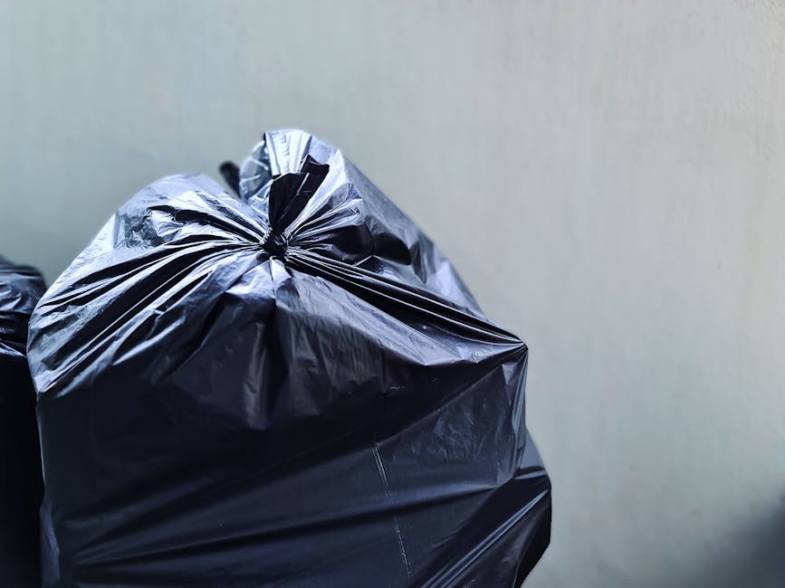 A collection of overflowing rubbish bins and scattered waste on a paved sidewalk in an urban area, including a grey mixed paper and card bin with its lid open, revealing crumpled paper, cardboard, and