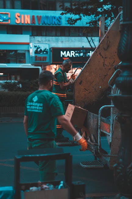 Two waste removal workers are operating a large industrial skip loader in an urban environment during twilight hours. The worker in the foreground wears a teal uniform with a logo and text on the back