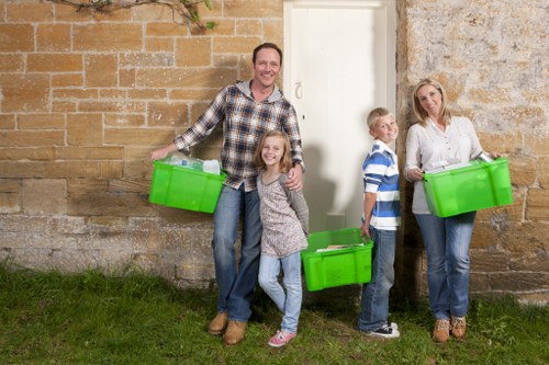 Four individuals, including a man, two children, and a woman, are standing outdoors on a grassy area beside a stone and brick wall. The man is smiling and holding a bright green plastic bin in his right hand, while a young girl stands in front of him, also smiling, with her arms around his waist. To the right, a boy and a woman each hold similar green bins; the boy is wearing a blue and white striped shirt and jeans, and the woman is dressed in a light blouse and jeans. The group appears to be engaged in a rubbish removal activity, with the green bins prominently displayed, set against a background of weathered brick and stonework. The scene suggests a community or family effort involving waste collection or clearance near a residential or commercial building, with natural daylight illuminating the setting. The presence of multiple rubbish bins and the casual outdoor environment aligns with services offered by Office Clearance Richmond for efficient rubbish removal in the Richmond area.