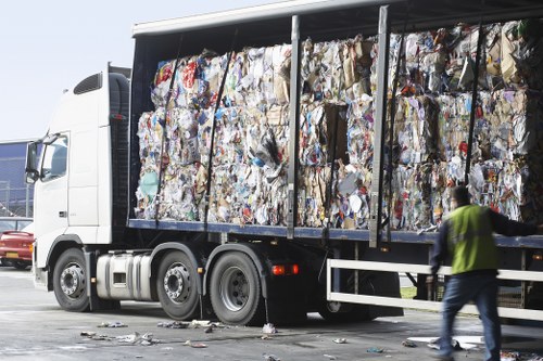 A large commercial lorry with a white cab and an open-sided trailer filled with compressed and crumpled mixed waste paper and cardboard, exhibiting various colours and textures, including brown kraft paper, printed packaging materials, and white office paper. The trailer is parked on a paved surface, possibly a busy street or industrial area near Richmond, with small scattered debris on the ground around the vehicle. In the foreground, a person wearing a high-visibility yellow vest and jeans is walking past the truck, slightly blurred due to motion. The background shows parts of other vehicles and a clear sky, suggesting daytime lighting. This image visually represents a professional rubbish removal or office clearance service, aligning with the offerings of Office Clearance Richmond in the local area.